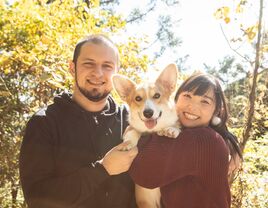 A couple smile, on a walk in the woods with their dog.