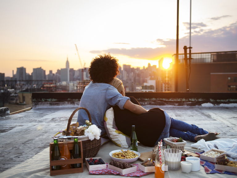 Couple embrace while watching a sunset at a rooftop picnic. 