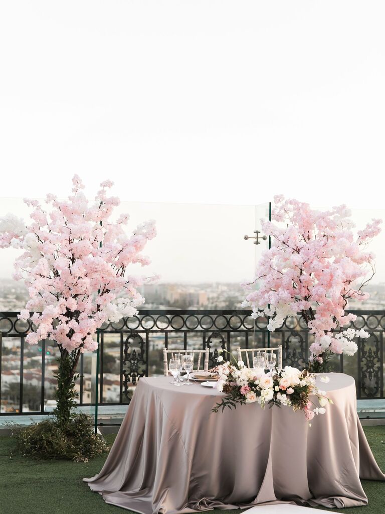outdoor wedding sweetheart table at rooftop venue with pink cherry blossom trees on either side and city view in the background