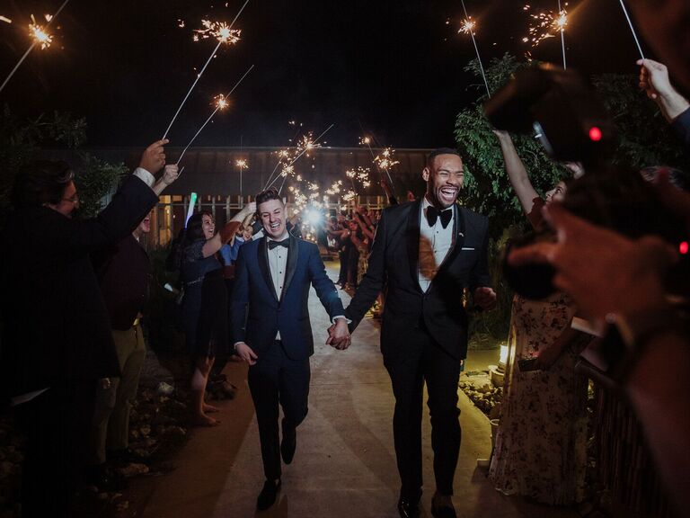 two grooms hold hands while walking through a tunnel of guests holding sparklers