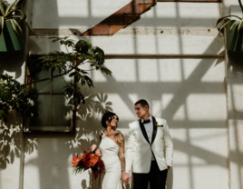 Bride and groom posing in front of white wall surrounded by plants