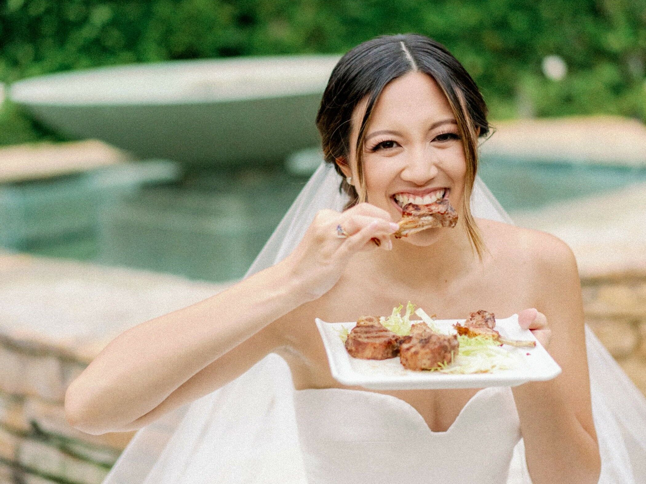 Bride smiles at the camera while eating chicken. 