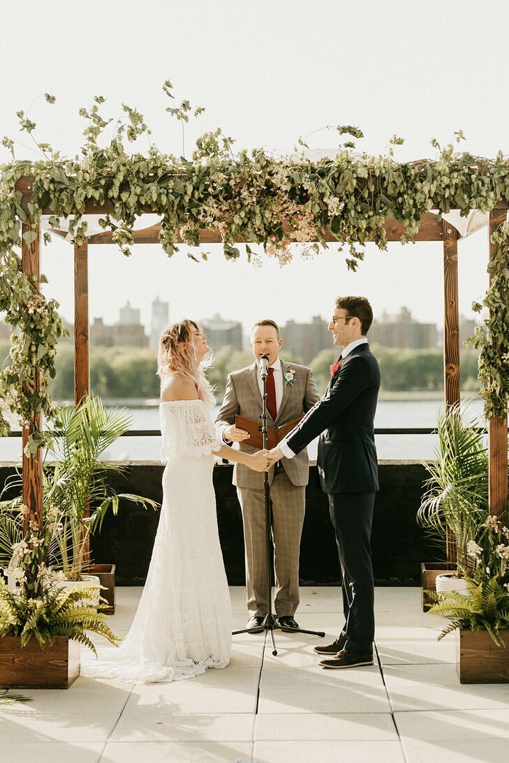 Couple holding hands under wood chuppah decorated with greenery.