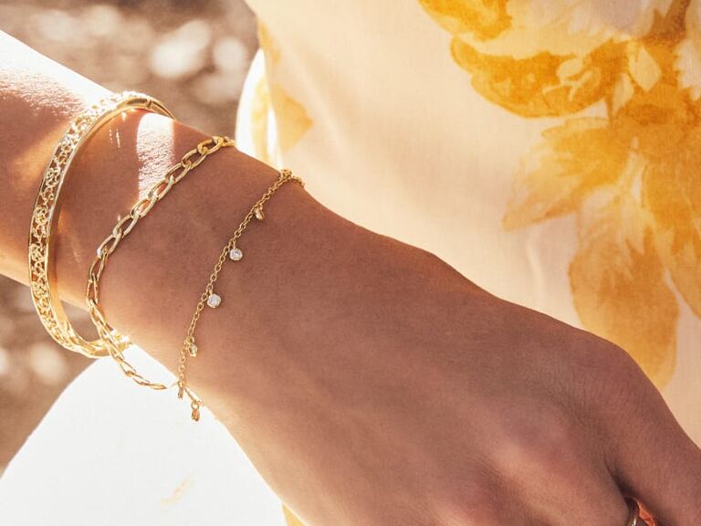 A set of three gold bracelets consisting of a herringbone bangle, a paperclip bracelet, and a chain bracelet with gems from Kendra Scott
