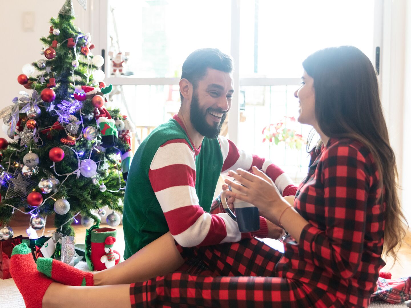 Young couple laughing and talking while enjoying a coffee wearing their christmas pajamas and sitting next to the christmas tree
