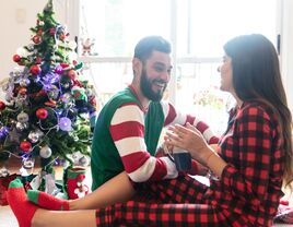 Young couple laughing and talking while enjoying a coffee wearing their christmas pajamas and sitting next to the christmas tree