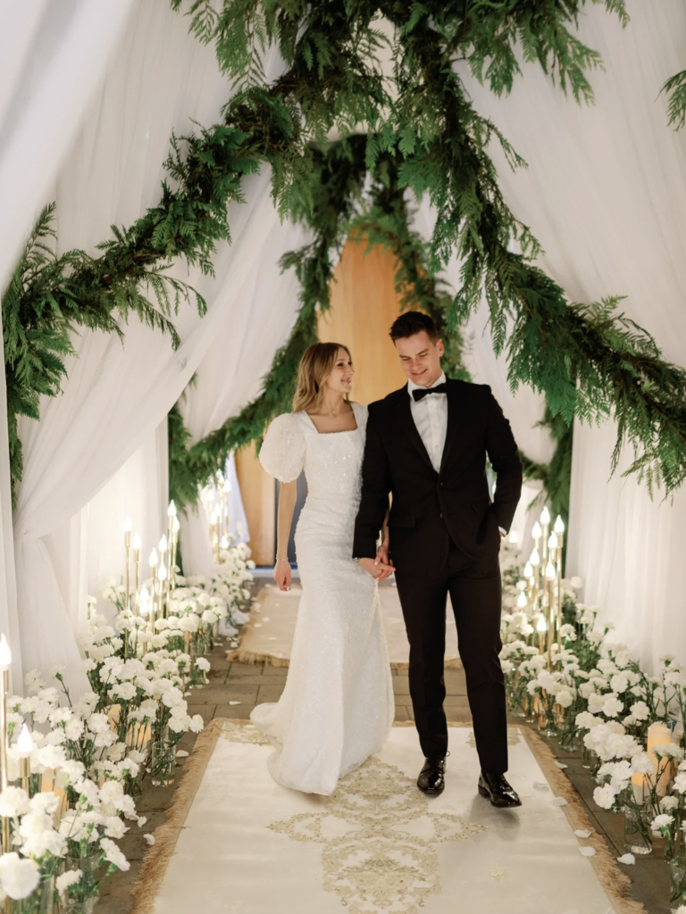 Groom in Tuxedo, Bride in Puff-Sleeved Gown Walk Under Draping, Hanging Greenery Entrance