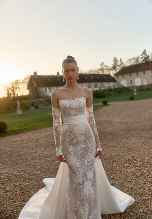 A structured Mikado and Chantilly lace bodice defines this Maison Blanche look. Floral motifs and a structured belt mark the natural waist, flaring into a trumpet silhouette. The strapless neckline shifts from straight to a gentle sweetheart curve, balancing an open low back. A separate light ivory overskirt transforms the profile into an A-line shape with a chapel train. Removable long sleeves finish this WONÃ Concept design.