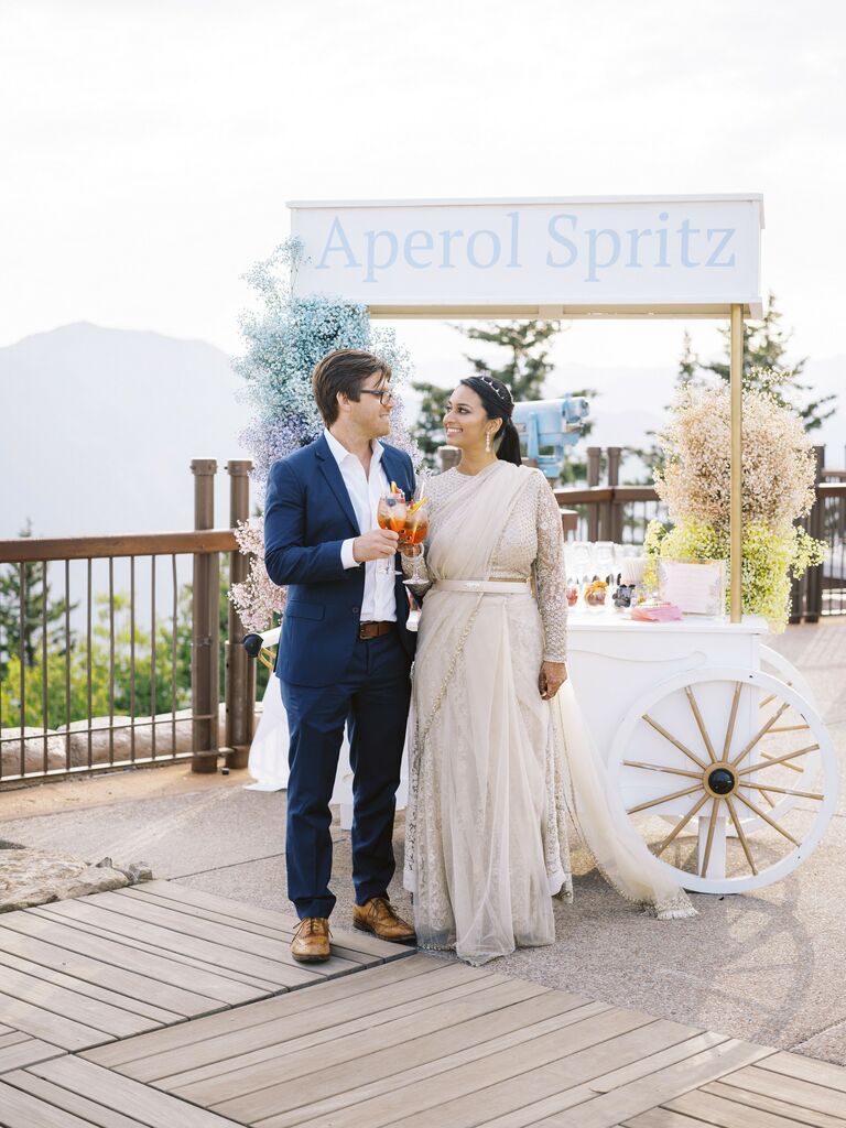 bride and groom standing in front of cocktail cart with sign that says aperol spritz