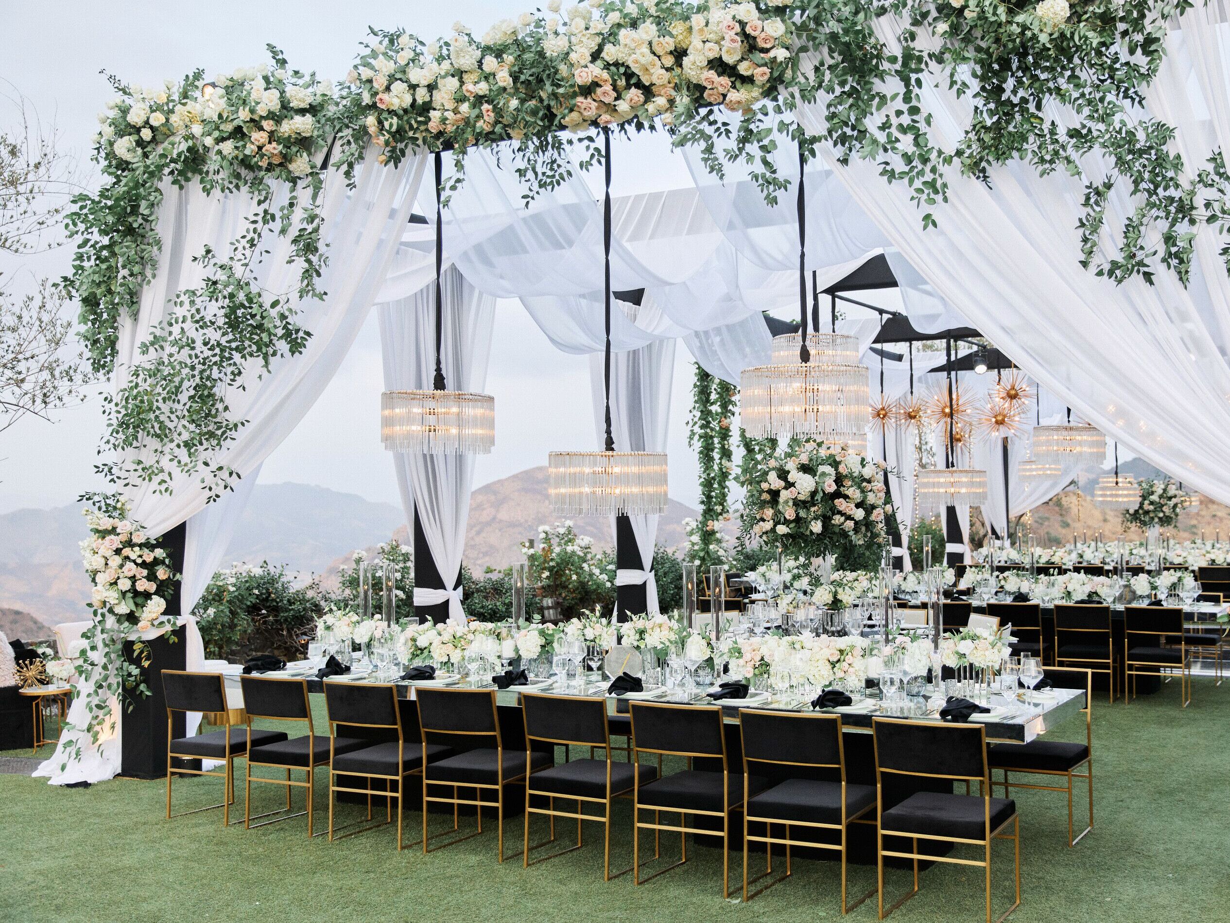 luxury outdoor wedding reception with white fabric draped across square canopies decorated with chandeliers, greenery and roses