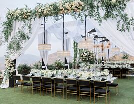 luxury outdoor wedding reception with white fabric draped across square canopies decorated with chandeliers, greenery and roses