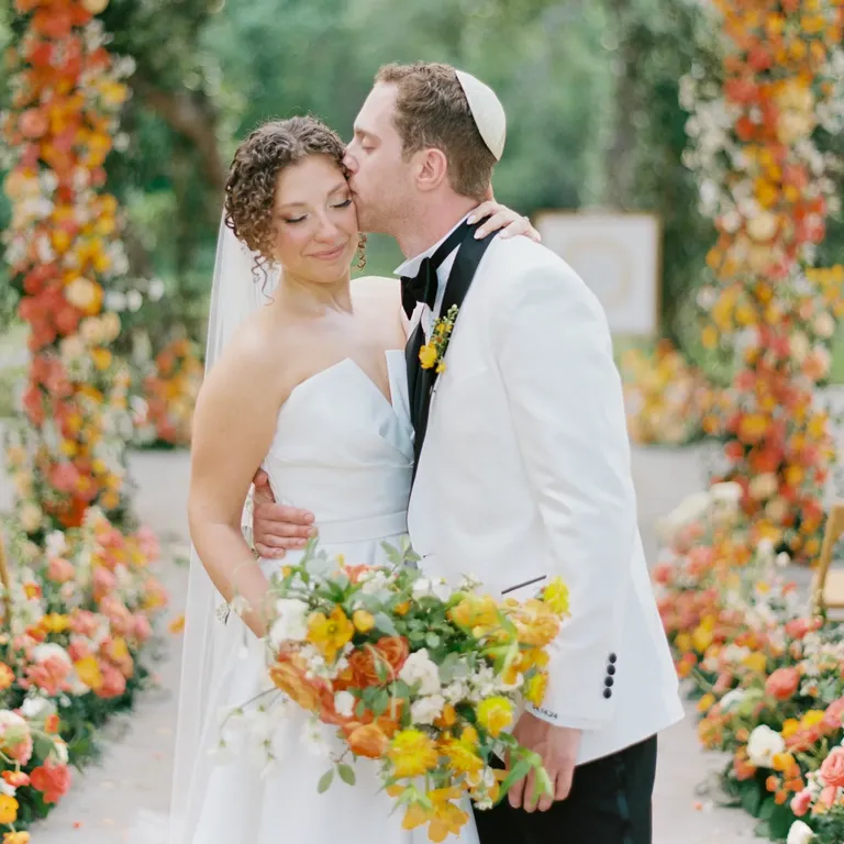 Groom kissing bride in front of orang and yellow Chuppah