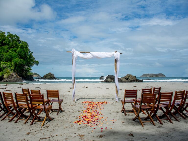 A wedding arch, rose petals and wood chairs sit ready on the beach to welcome the happy couple and their guests in Costa Rica.