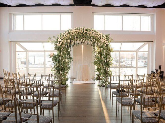 Indoor ceremony site with a foliage arch and large windows
