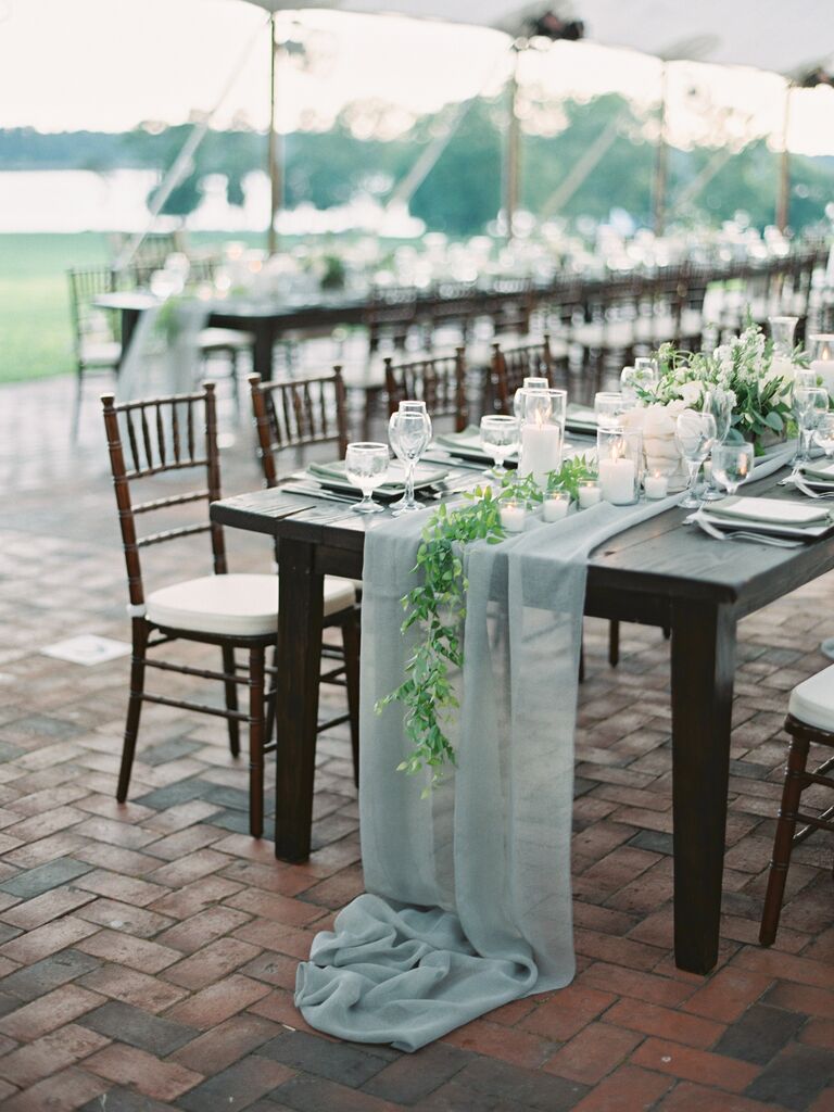 simple wedding centerpiece with long dark wood banquet table, sheer light blue runner, greenery and white votive candles