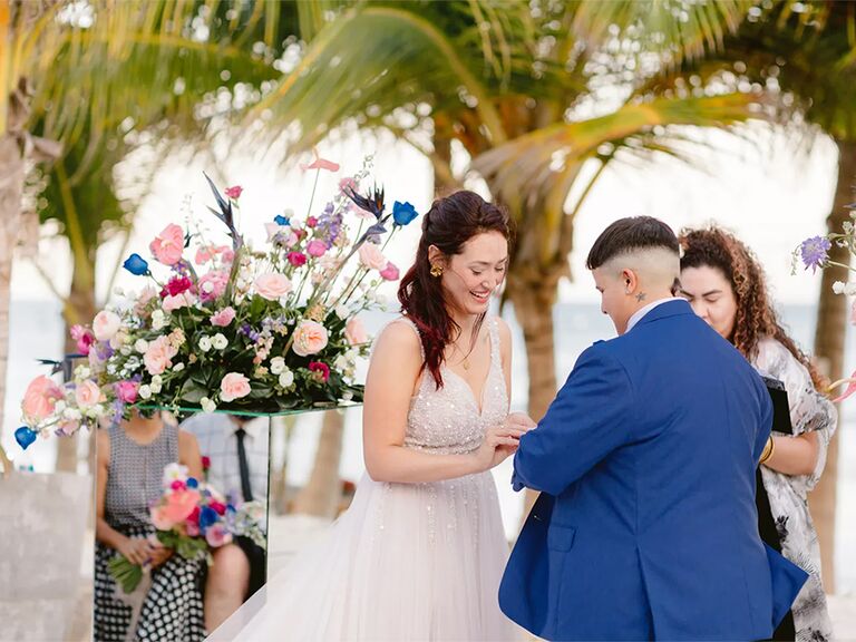 Couple exchanging rings at beachfront ceremony with colorful bouquets