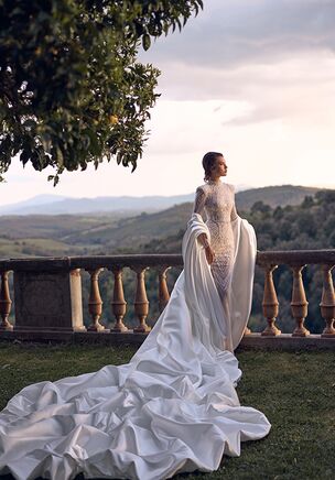 Aphrodite gown is the true showpiece of the collection – a masterpiece of elegance and drama. This mermaid silhouette lace dress features a high neckline and long fitted sleeves that fasten with invisible zippers, offering both sophistication and comfort. Fully adorned with intricate lace patterns and delicate embroidery, the gown exudes timeless romance. The look is elevated by a spectacular detachable three-meter cape made of shimmering mikado, which gracefully drapes and creates a majestic layered effect. The cape loops over the forearm, forming a voluminous silhouette that flows into a grand train. The back of the dress closes with a concealed zipper and includes a subtle train for a refined finish. Aphrodite is designed for brides who dream of regal beauty and unforgettable entrances.