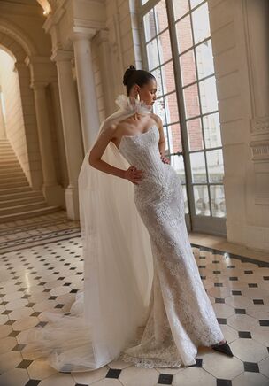 The Norma bridal set introduces a visual transformation centered around a sleek gown tailored from textured textile and rich lace. The fitted bodice features a dipped, classic neckline before widening below the hips with a dropped waist, releasing into a court-length train. For an alternate presentation, a detachable ivory cape rests over bare shoulders, adding soft volume past the hemline. This layered design allows alternate styling. Delicate floral textures cascade down the surface, adding tactile dimension without interrupting clean lines. The open back balances the front coverage, creating a striking rear view as the cape drapes over the dress.
