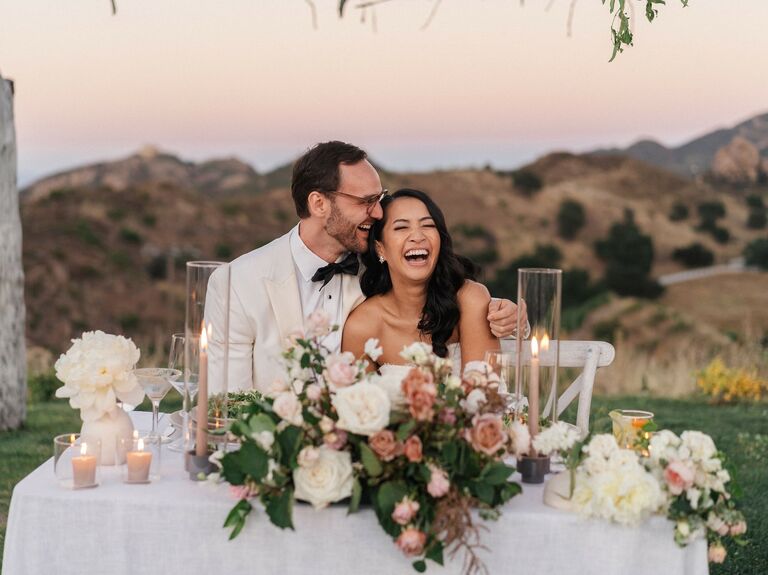 bride and groom laugh while listening to toast seated at outdoor sweetheart table decorated with light pink rose centerpiece, taper candles and mountain scenery in the background 