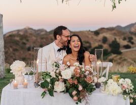 bride and groom laugh while listening to toast seated at outdoor sweetheart table decorated with light pink rose centerpiece, taper candles and mountain scenery in the background 