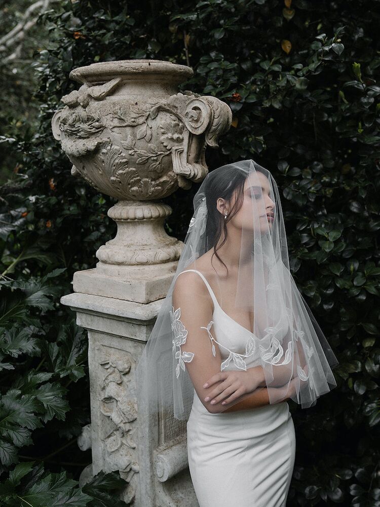 Model stands in a garden and wears a sheer veil over her face. 