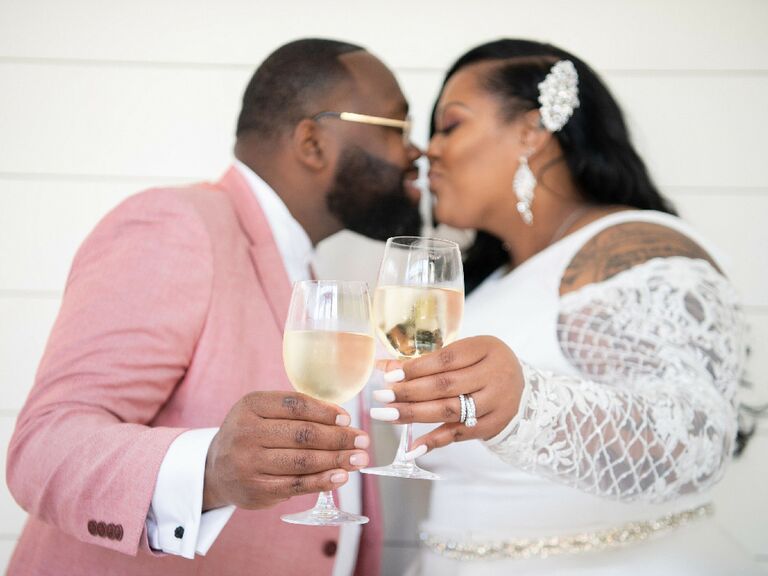 Groom wearing pink suit kissing bride while holding glass of wine