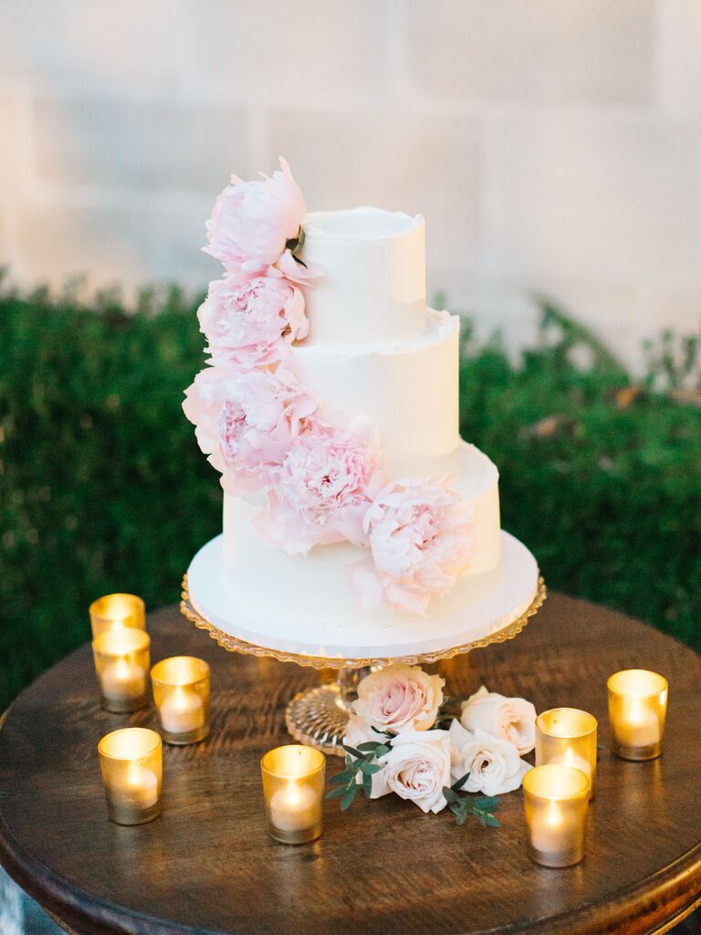 three tier buttercream wedding cake decorated with pink peonies on wooden table surrounded by votive candles
