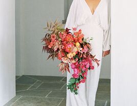 bride wearing white wedding jumpsuit holds cascading bouquet with orange roses, coral peonies and pink bougainvillea