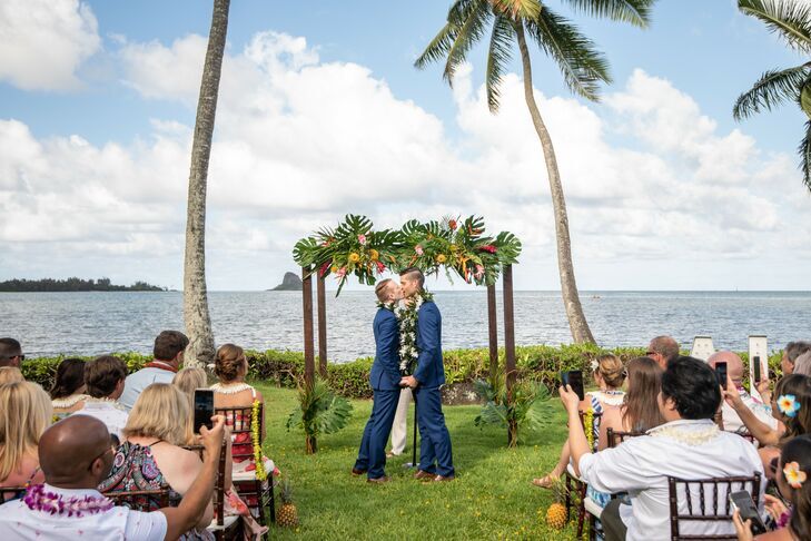 Grooms sharing first kiss in front of tropical chuppah at beach wedding.