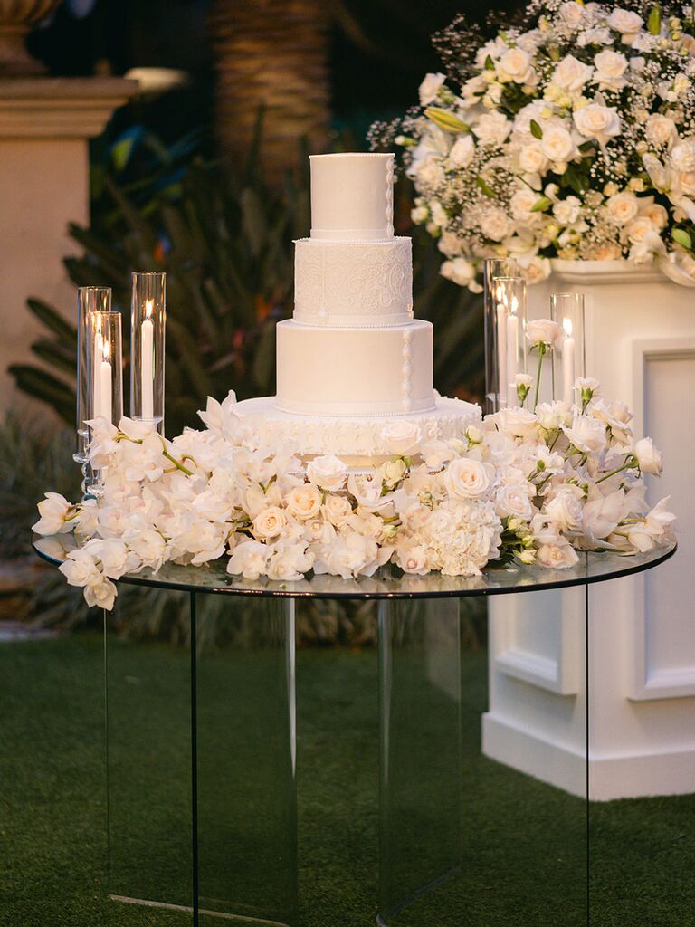 2024 wedding cake decorated with white fondant surrounded by white flowers and taper candles on a glass table