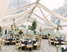 Partially draped clear event tent with round tables, gold chairs and hanging foliage