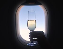 Close-up of silhouette hand holding champagne glass against airplane window during flight on sunny day.