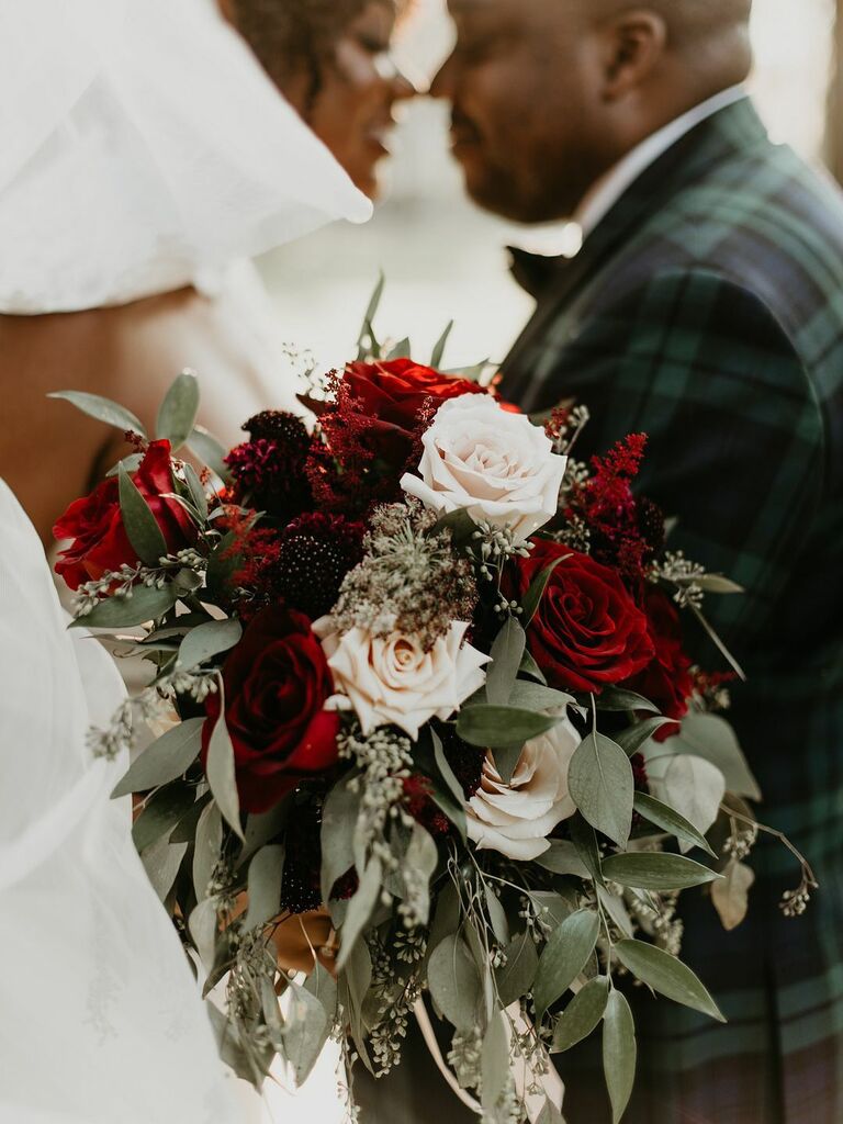 closeup of winter wedding bouquet featuring light pink roses, red roses and greenery with couple kissing in background