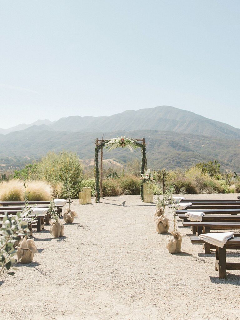 outdoor wedding ceremony with long benches and potted trees as aisle markers