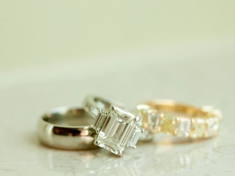 close-up of three-stone emerald cut engagement ring on silver band with yellow gold diamond wedding band and groom's silver wedding band