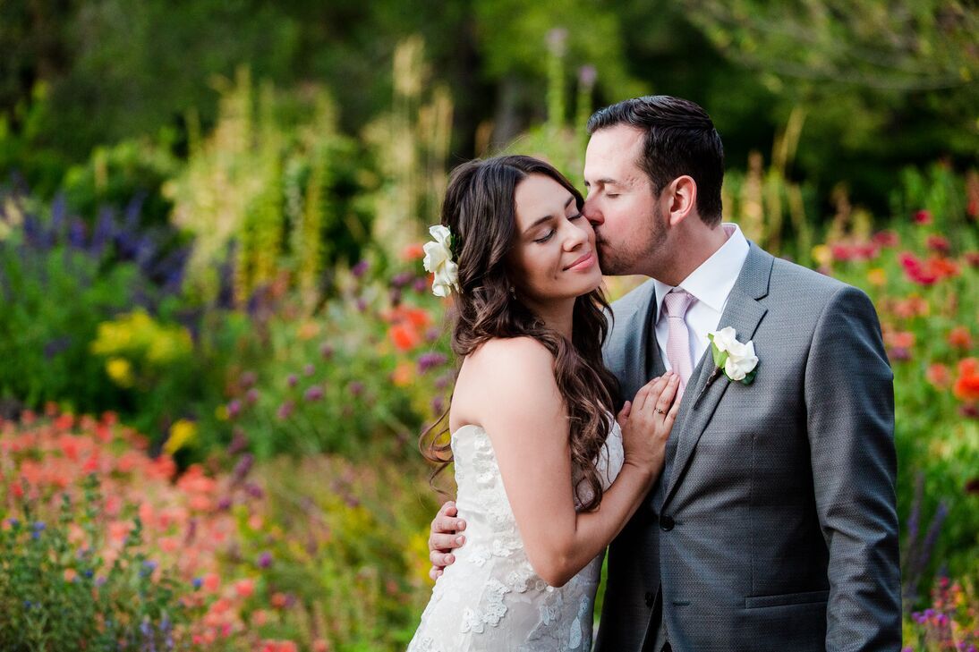 Newlyweds share a sweet kiss surrorunded by colorful flowers in a lush garden. 