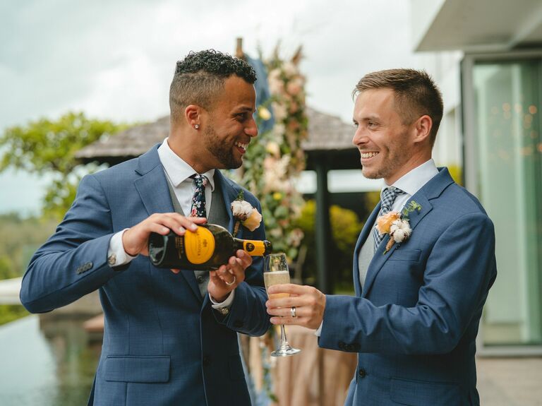 two grooms smile and pour champagne wearing navy blue suits