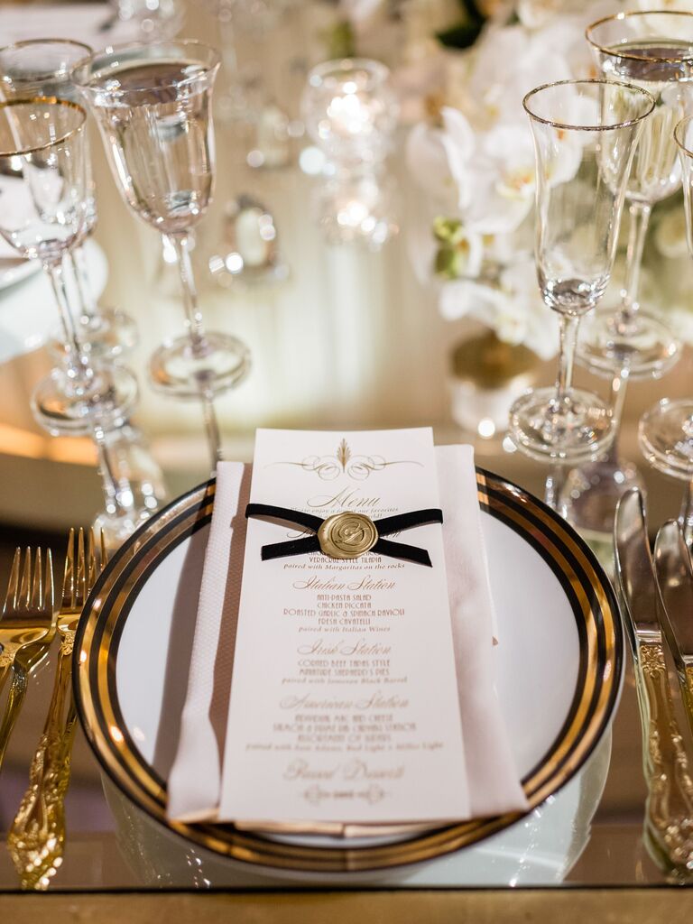 wedding menu card decorated with black ribbon and gold wax seal on top of gold-rimmed dinner plate