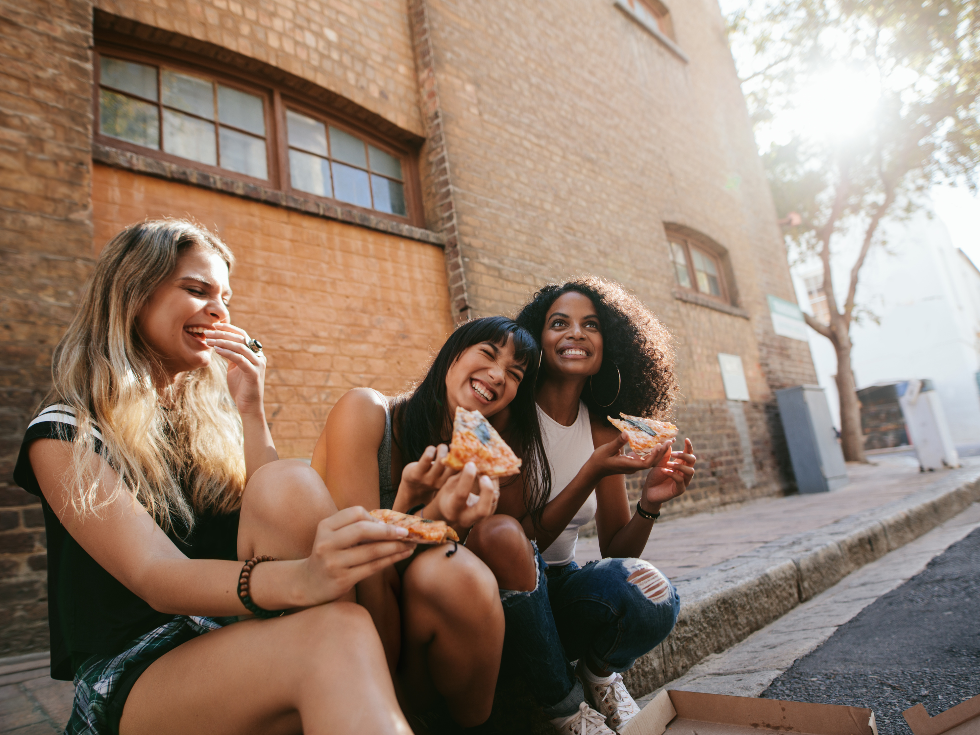 Group of freinds sitting on sidewalk eating pizza