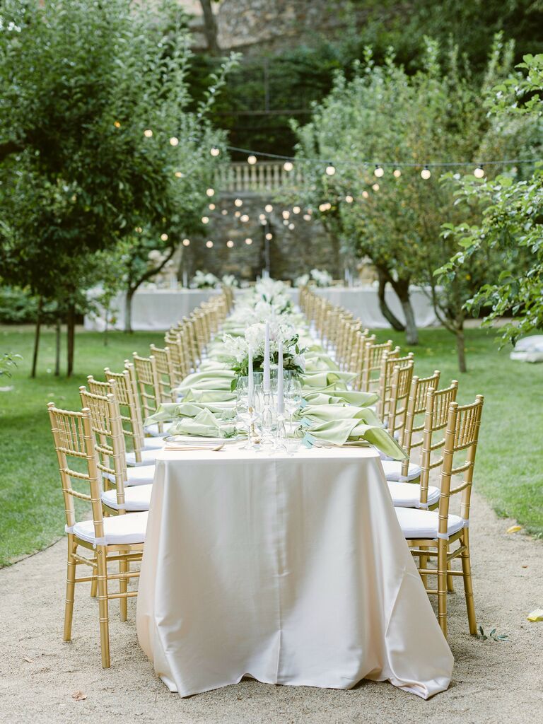 outdoor wedding reception with long banquet table decorated with iridescent cream table linens, taper candles and gold chiavari chairs