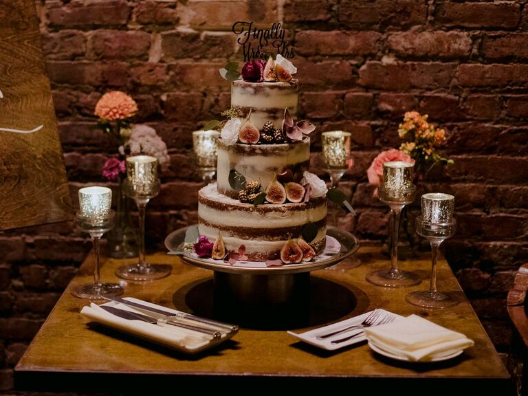 three tier semi naked wedding cake decorated with sliced figs sits on a wooden table against a brick wall