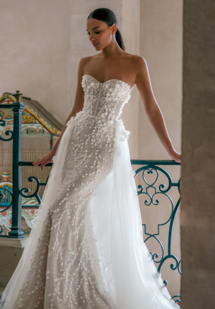 Perfectly flattering the figure and talking magically to a woman's curves it takes up to 90 hours of handwork to create this dress. Jazz is a sheath feminine wedding gown with a lace strapless top. The corset is embellished with sparkles and fine 3D floral lace. Lace appliques elevate the inherent texture of the Chantilly lace down the skirt that is fitted to perfection. Accessorized with a butter soft Tulle overskirt with floral appliques near the bride's waistline it takes on extra volume for a grand down-the-aisle entrance.