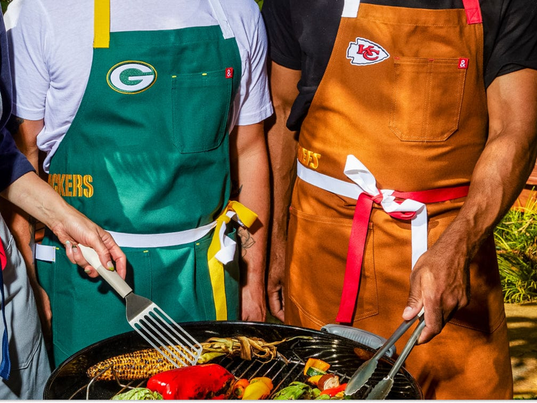 Hedley & Bennett professional aprons embroidered with the sports' teams Greenbay Packers and Kansas City Chiefs two men wearing them around a grill