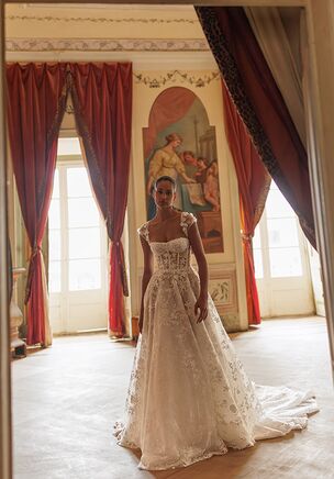 A romantic A-line silhouette gown crafted from delicate lace and adorned with 3D floral applications. The bodice cups are softly draped with tulle, creating a light and feminine look, while the straps are embellished with intricate floral details. The straps are easily removable, allowing for a quick transformation of the style. The dress features a lace-up corset for a secure and flattering fit, and a long, flowing train that adds a graceful and dramatic finish.