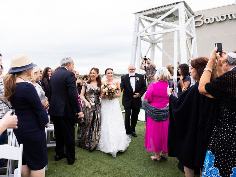 A bride and her parents walk down the aisle at an outdoor ceremony.