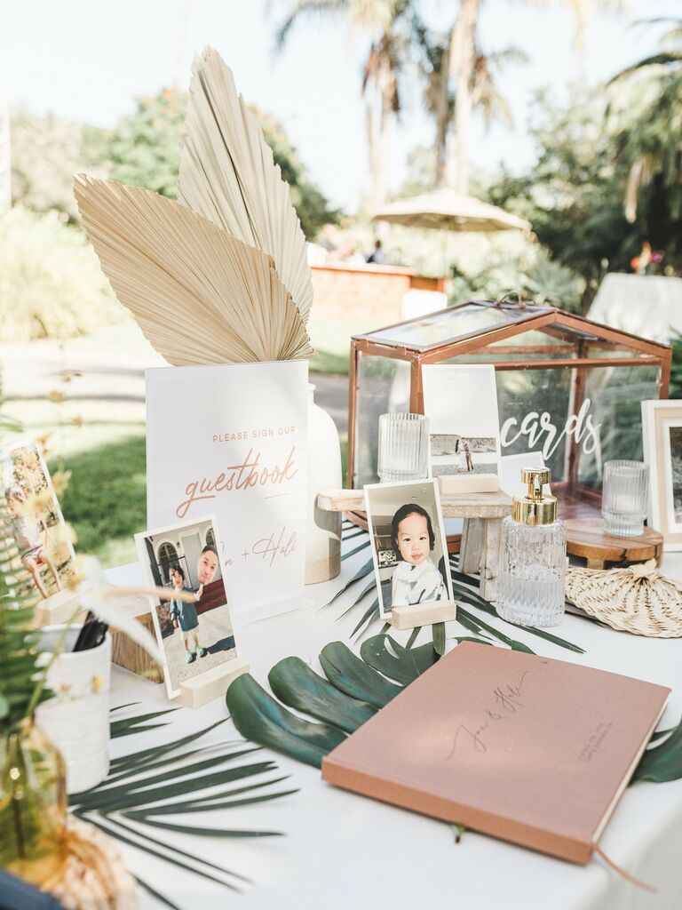 wedding welcome table decorated with gold glass card box, childhood photos of couple, tropical greenery and dried palm leaves