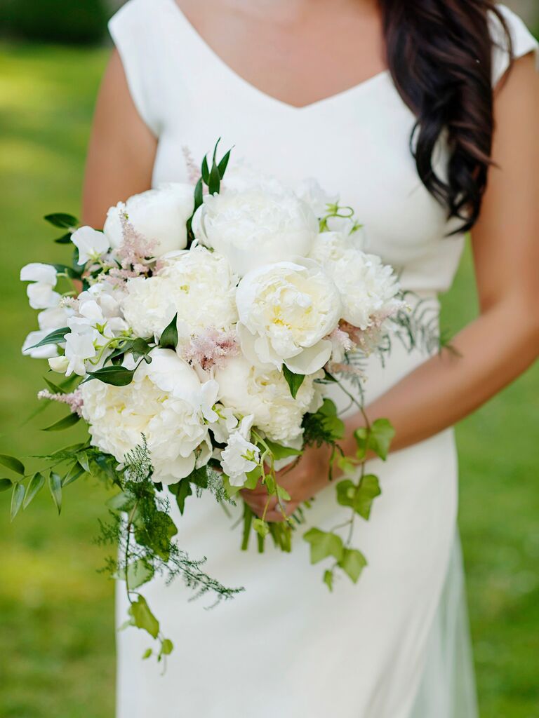 A bride stands holding a burgeoning white peony bouquet.