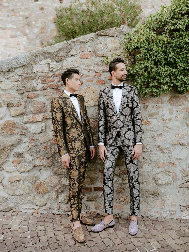 two grooms stand against a stone wall wearing metallic silver and gold brocade pattern tuxedos