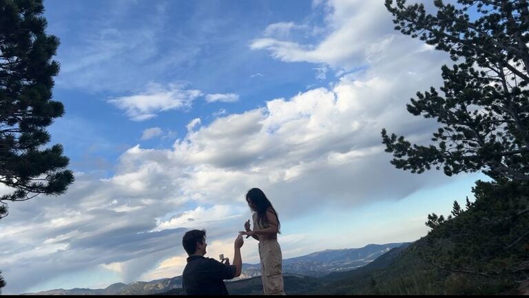 Will proposed to Magi in beautiful Estes Park, Colorado because she loves the mountains and nature. 
The day before, he told her they were going to go on a date but didn't say where. This picture is a screenshot of  video taken during the proposal. When we got to the place Will wanted to propose, he told me "This would be a beautiful landscape for a picture." I didn't know he was recording.
I turned and said, "You are right--it is a beautiful landscape" without looking at him and when I turned around, he was down on one knee.  He said:  "Magi Junco, I love you so much, would you marry me?" I started crying of happiness.
Will confessed later that he was worried about losing the ring during the trip because we had to walk quite far to get to the place in the photo where he proposed.


