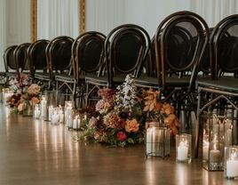 indoor wedding ceremony aisle with dark wooden chairs, white candles in glass vases and orange, white and burgundy flower arrangements on the floor